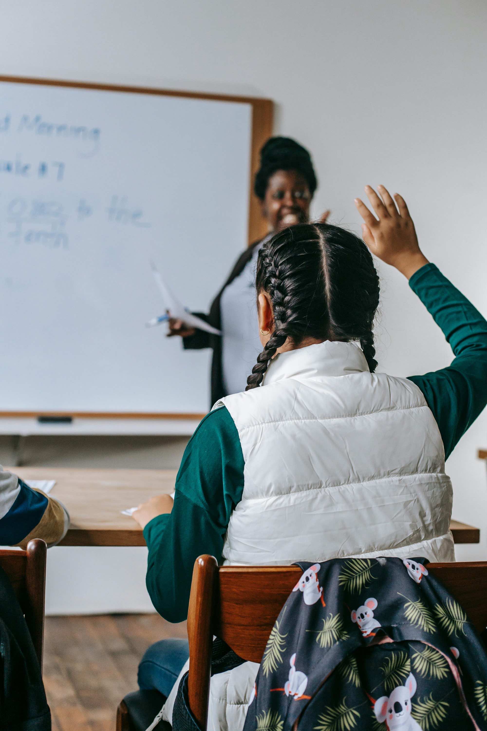 Student raising hand while teacher leads class at whiteboard